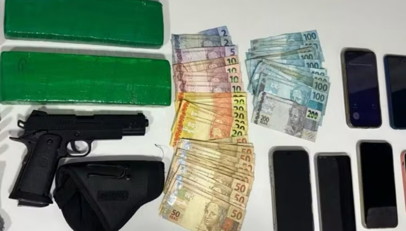 Assorted items on a table two large green wrapped bundles a black handgun with a holster and stacks of Brazilian real banknotes next to multiple mobile phones