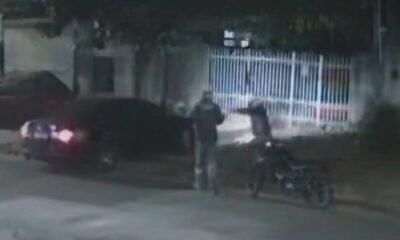 Two people stand beside a motorcycle at night near a parked car and a white metal fence