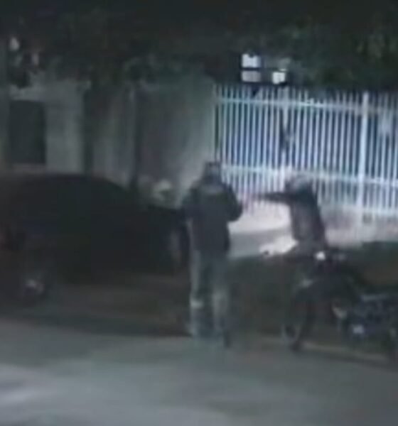 Two people stand beside a motorcycle at night near a parked car and a white metal fence