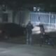 Two people stand beside a motorcycle at night near a parked car and a white metal fence