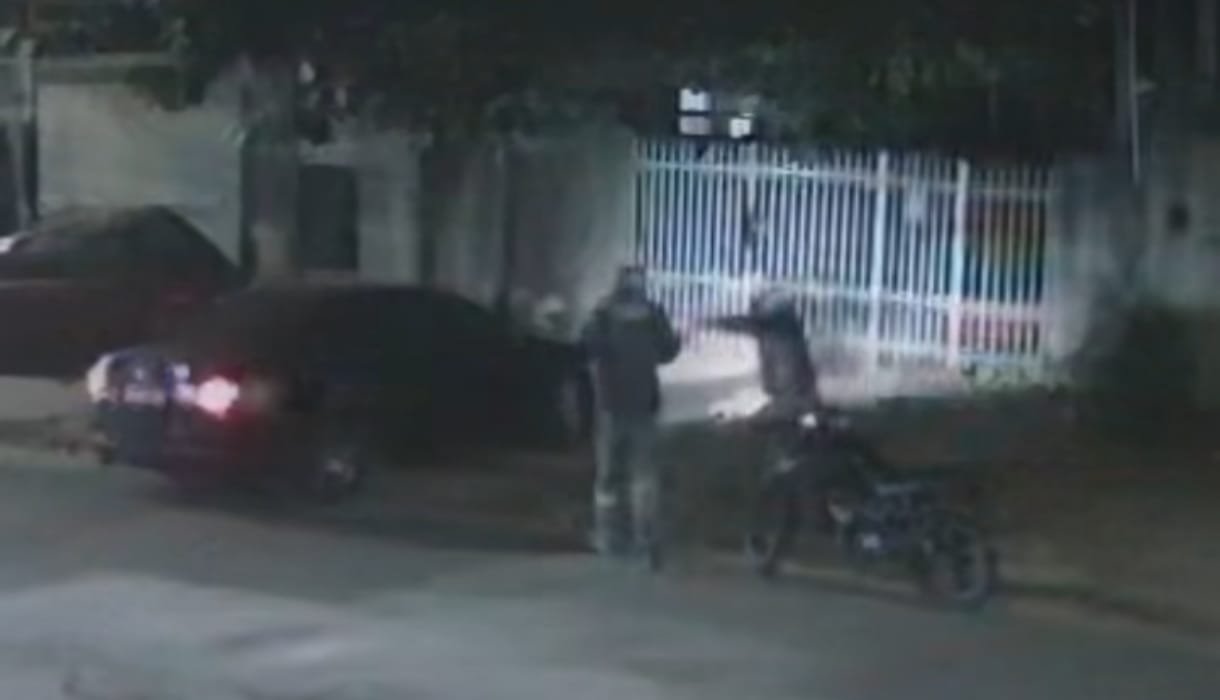 Two people stand beside a motorcycle at night near a parked car and a white metal fence