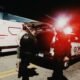 Nighttime emergency scene with an ambulance and a GCM police vehicle responders near the open trunk with red flashing lights in the background