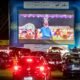 Drive in cinema at night with a large blue inflatable screen frame and a big cartoon movie on screen cars lined up in front watching the show