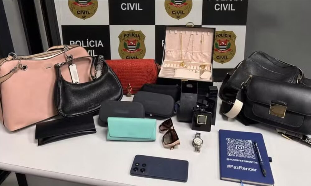 Display of seized items on a table: pink and black handbags, purses, a red clutch, wallets, sunglasses, a wristwatch, jewelry box, perfume bottle, and a smartphone against a police backdrop.