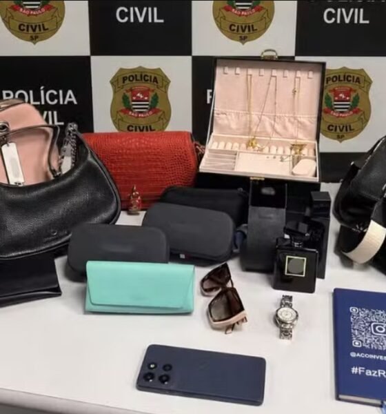 Display of seized items on a table pink and black handbags purses a red clutch wallets sunglasses a wristwatch jewelry box perfume bottle and a smartphone against a police backdrop