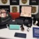 Display of seized items on a table pink and black handbags purses a red clutch wallets sunglasses a wristwatch jewelry box perfume bottle and a smartphone against a police backdrop