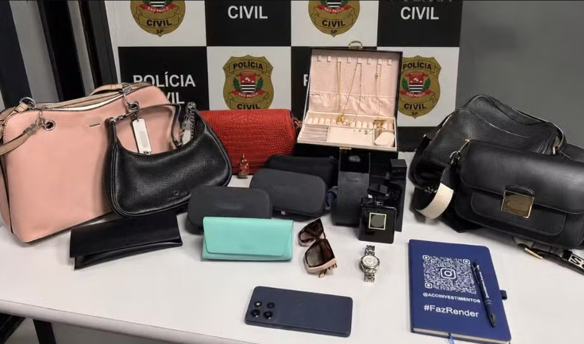 Display of seized items on a table pink and black handbags purses a red clutch wallets sunglasses a wristwatch jewelry box perfume bottle and a smartphone against a police backdrop