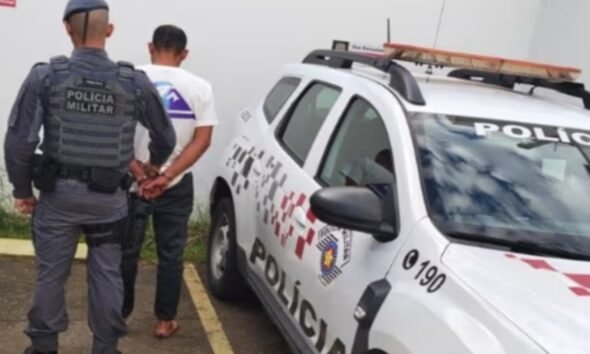 Military police officer cuffs a man next to a white police SUV with a rooftop light bar in a parking area