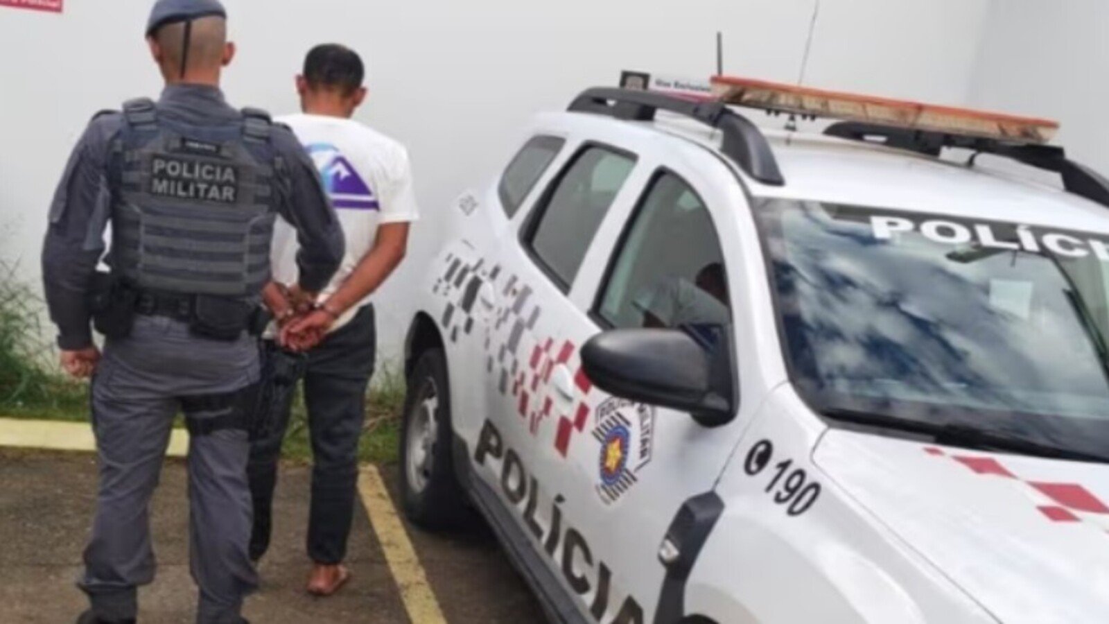 Military police officer cuffs a man next to a white police SUV with a rooftop light bar in a parking area