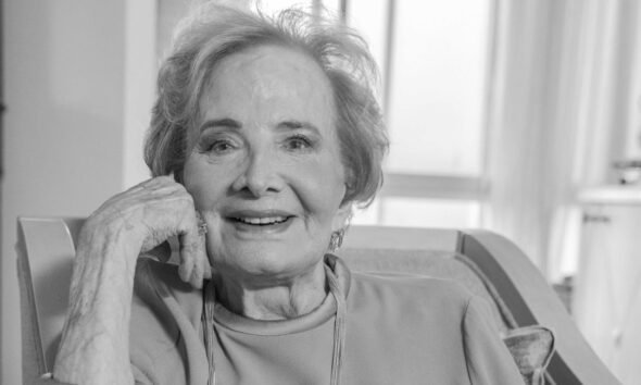 Smiling elderly woman seated in a chair resting her chin on her hand looking at the camera in a cozy home setting