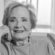 Smiling elderly woman seated in a chair resting her chin on her hand looking at the camera in a cozy home setting