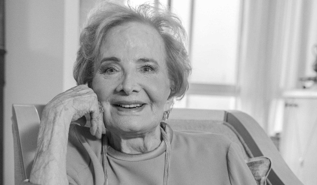 Smiling elderly woman seated in a chair resting her chin on her hand looking at the camera in a cozy home setting