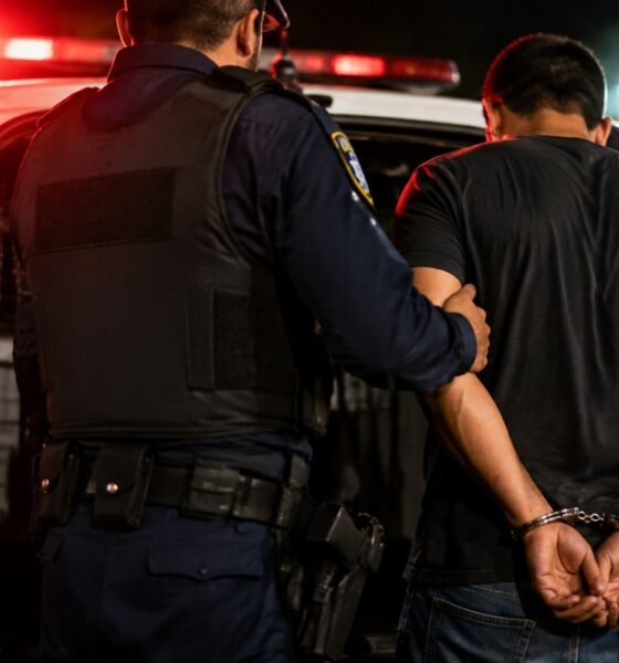 Officer in tactical vest guiding a handcuffed detainee toward the open police SUV at night red emergency lights flashing in the background