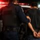 Officer in tactical vest guiding a handcuffed detainee toward the open police SUV at night red emergency lights flashing in the background