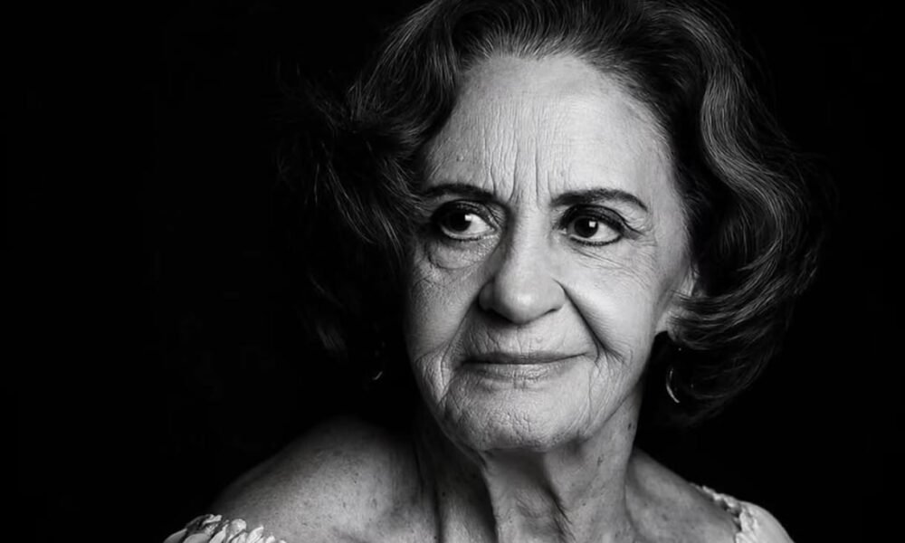 Black and white portrait of an elderly woman with short wavy hair looking to the left wearing a light off shoulder top against a dark background