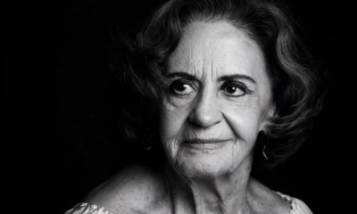 Black and white portrait of an elderly woman with short wavy hair looking to the left wearing a light off shoulder top against a dark background