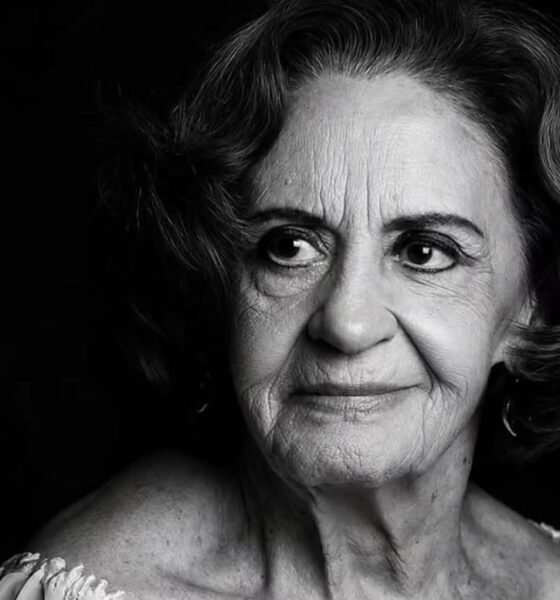 Black and white portrait of an elderly woman with short wavy hair looking to the left wearing a light off shoulder top against a dark background