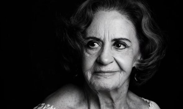 Black and white portrait of an elderly woman with short wavy hair looking to the left wearing a light off shoulder top against a dark background