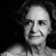 Black and white portrait of an elderly woman with short wavy hair looking to the left wearing a light off shoulder top against a dark background