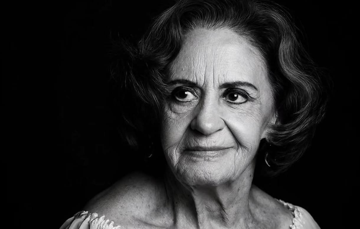 Black and white portrait of an elderly woman with short wavy hair looking to the left wearing a light off shoulder top against a dark background
