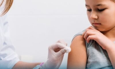 Child receives a vaccination in the upper arm from a gloved healthcare worker inside a clinical setting looking away calmly