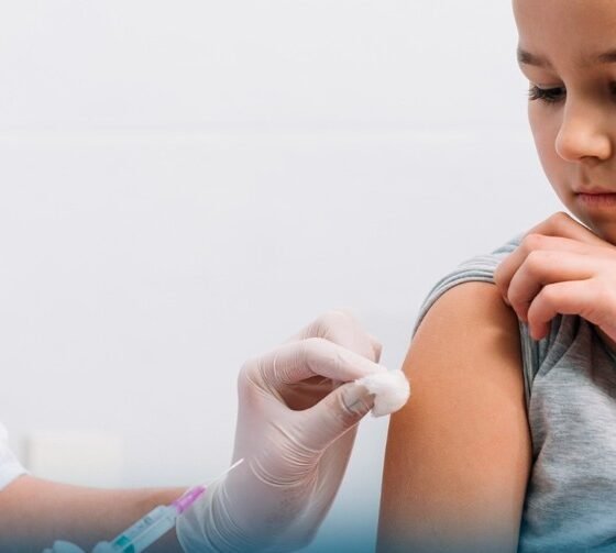 Child receives a vaccination in the upper arm from a gloved healthcare worker inside a clinical setting looking away calmly