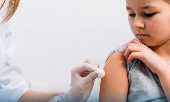 Child receives a vaccination in the upper arm from a gloved healthcare worker inside a clinical setting looking away calmly