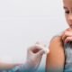 Child receives a vaccination in the upper arm from a gloved healthcare worker inside a clinical setting looking away calmly