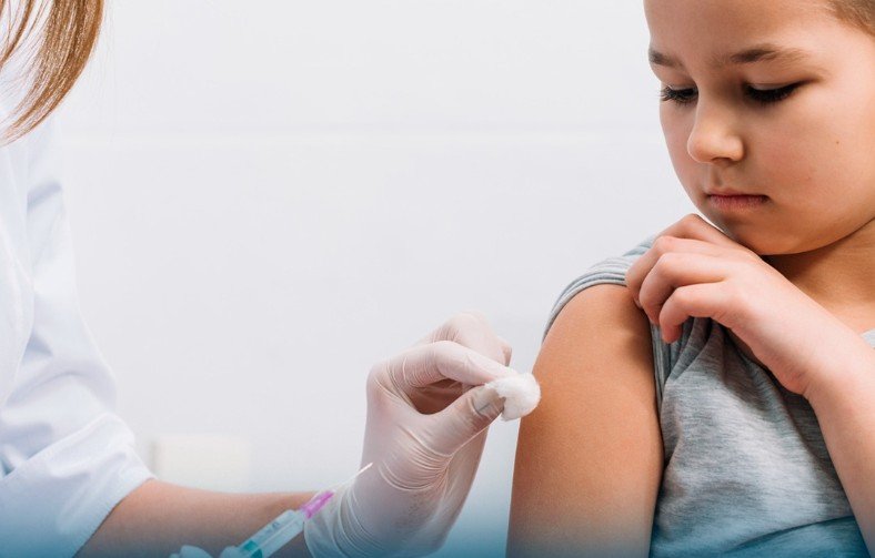 Child receives a vaccination in the upper arm from a gloved healthcare worker inside a clinical setting looking away calmly